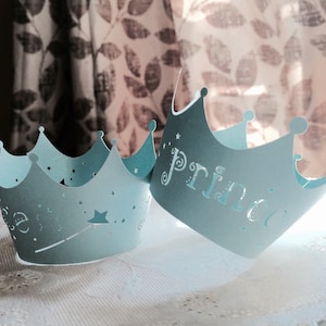 Puede incluir: Dos coronas de papel azul claro con un dise&ntilde;o de estrella y varita m&aacute;gica. Una corona dice "Pr&iacute;ncipe" y la otra dice "Princesa".