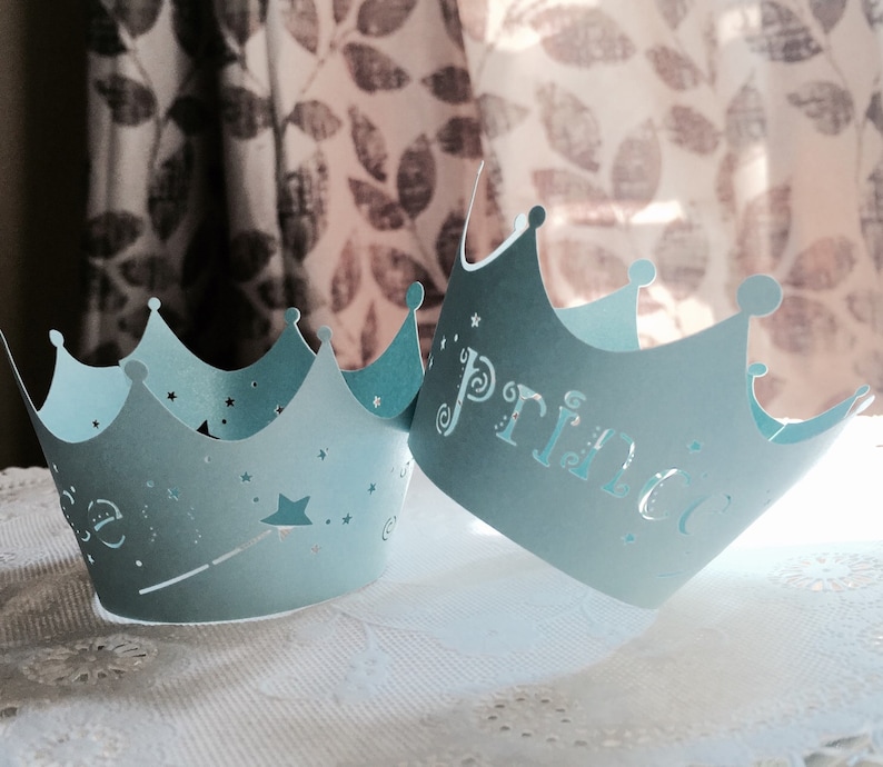 Puede incluir: Dos coronas de papel azul claro con un dise&ntilde;o de estrella y varita m&aacute;gica. Una corona dice "Pr&iacute;ncipe" y la otra dice "Princesa".