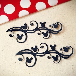 May include: Two black paper cutouts of a decorative floral design with Mickey Mouse heads. The design is intricate and features swirls and loops.