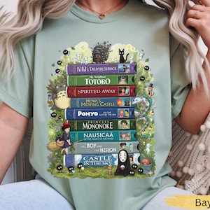 K&ouml;nnte beinhalten: Hellgr&uuml;nes T-Shirt mit einem Stapel illustrierter B&uuml;cher, darunter Titel wie "Kiki's Delivery Service" und "Mein Nachbar Totoro". Das Design zeigt Charaktere und Elemente aus verschiedenen Zeichentrickfilmen, umgeben von skurrilen Details.