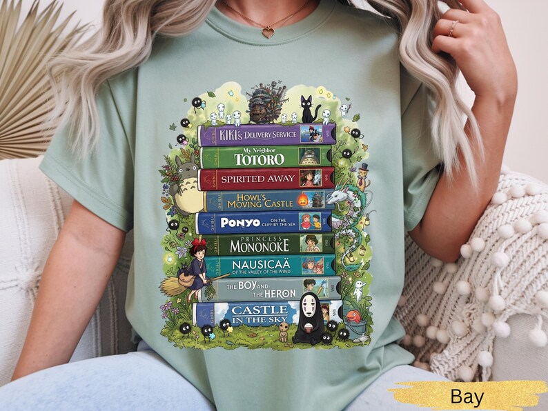 K&ouml;nnte beinhalten: Hellgr&uuml;nes T-Shirt mit einem Stapel illustrierter B&uuml;cher, darunter Titel wie "Kiki's Delivery Service" und "Mein Nachbar Totoro". Das Design zeigt Charaktere und Elemente aus verschiedenen Zeichentrickfilmen, umgeben von skurrilen Details.