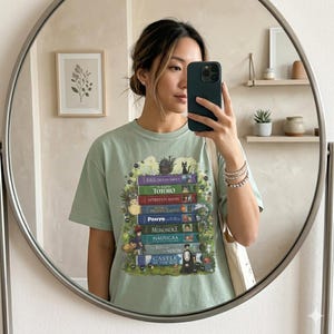 K&ouml;nnte beinhalten: Hellgr&uuml;nes T-Shirt mit einem Stapel B&uuml;cher mit Titeln wie "Totoro" und "Spirited Away". Das Design enth&auml;lt Illustrationen von Charakteren aus verschiedenen Zeichentrickfilmen. Die Person im Bild tr&auml;gt das T-Shirt.