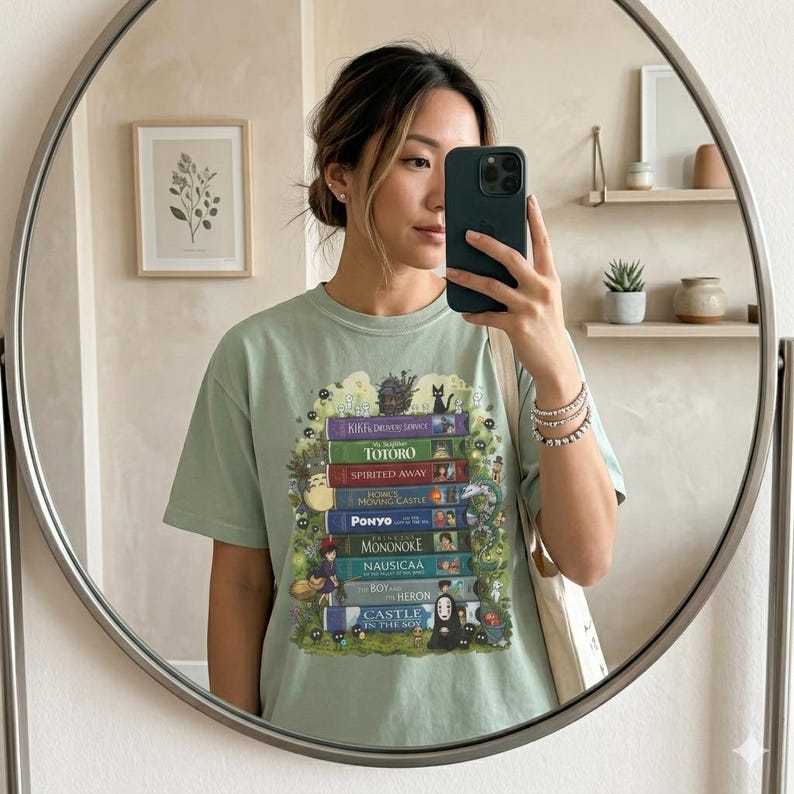 K&ouml;nnte beinhalten: Hellgr&uuml;nes T-Shirt mit einem Stapel B&uuml;cher mit Titeln wie "Totoro" und "Spirited Away". Das Design enth&auml;lt Illustrationen von Charakteren aus verschiedenen Zeichentrickfilmen. Die Person im Bild tr&auml;gt das T-Shirt.