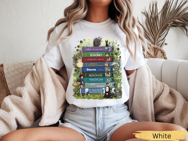 K&ouml;nnte beinhalten: Wei&szlig;es T-Shirt mit einem farbenfrohen Grafikdesign von gestapelten B&uuml;chern mit Titeln wie "Totoro" und "Spirited Away". Das Design enth&auml;lt Illustrationen von Charakteren und Bl&auml;ttern, was einen skurrilen und k&uuml;nstlerischen Look erzeugt. Das Wort "White" steht unten.
