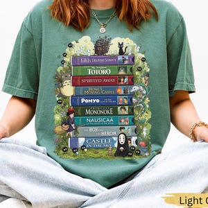 K&ouml;nnte beinhalten: Hellgr&uuml;nes T-Shirt mit einem Design aus gestapelten B&uuml;chern, die mit animierten Filmtiteln und Charakteren versehen sind. Zu den Titeln geh&ouml;ren "Kikis kleiner Lieferservice", "Mein Nachbar Totoro" und "Chihiros Reise ins Zauberland".