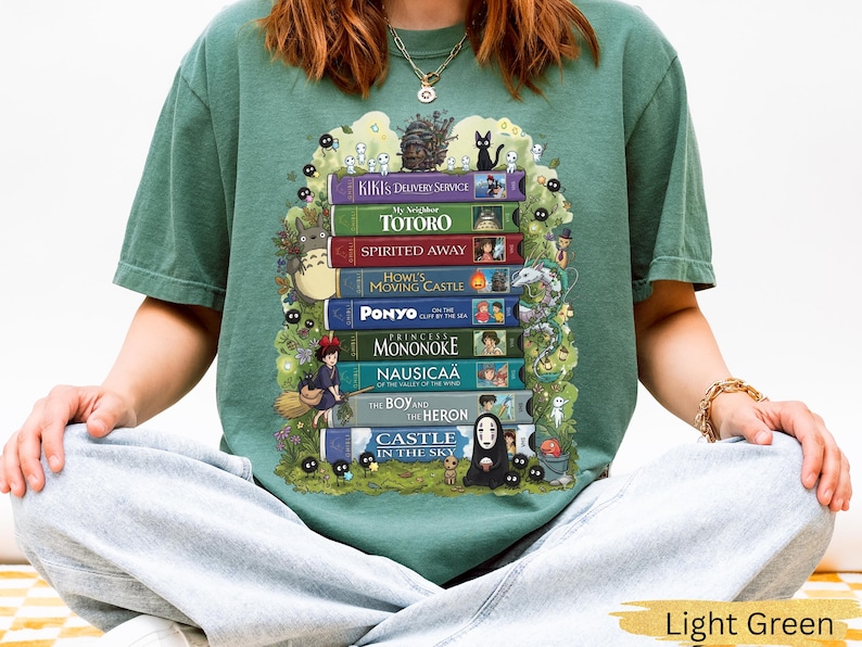 K&ouml;nnte beinhalten: Hellgr&uuml;nes T-Shirt mit einem Design aus gestapelten B&uuml;chern, die mit animierten Filmtiteln und Charakteren versehen sind. Zu den Titeln geh&ouml;ren "Kikis kleiner Lieferservice", "Mein Nachbar Totoro" und "Chihiros Reise ins Zauberland".
