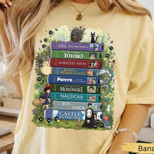 K&ouml;nnte beinhalten: Hellgelbes T-Shirt mit einem Aufdruck von gestapelten B&uuml;chern, die mit Titeln wie "Chihiros Reise ins Zauberland" und "Mein Nachbar Totoro" versehen sind. Das Design enth&auml;lt Charaktere aus verschiedenen Filmen. Das Wort "Banana" ist unten aufgedruckt.