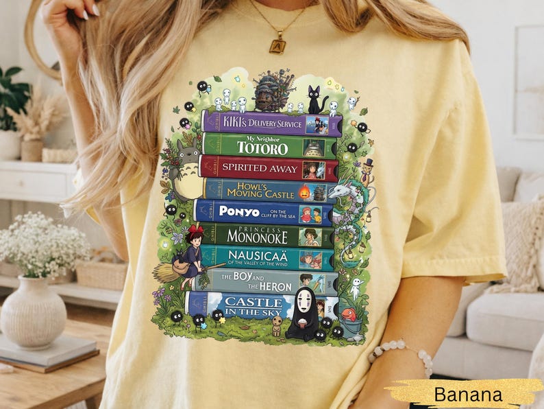 K&ouml;nnte beinhalten: Hellgelbes T-Shirt mit einem Aufdruck von gestapelten B&uuml;chern, die mit Titeln wie "Chihiros Reise ins Zauberland" und "Mein Nachbar Totoro" versehen sind. Das Design enth&auml;lt Charaktere aus verschiedenen Filmen. Das Wort "Banana" ist unten aufgedruckt.