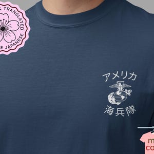 May include: A navy blue t-shirt featuring a white graphic on the left chest. The graphic displays the United States Marine Corps emblem with the Japanese text "アメリカ" above and "海兵隊" below. A pink circular badge with a cherry blossom icon is in the top left corner.