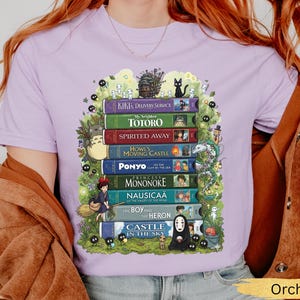 K&ouml;nnte beinhalten: Orchideenfarbenes T-Shirt mit einem Design aus bunten B&uuml;chern, darunter Titel wie "Kiki's Delivery Service" und "Mein Nachbar Totoro." Das Design enth&auml;lt Illustrationen von Charakteren und Elementen aus den Filmen, umgeben von skurrilen Details.