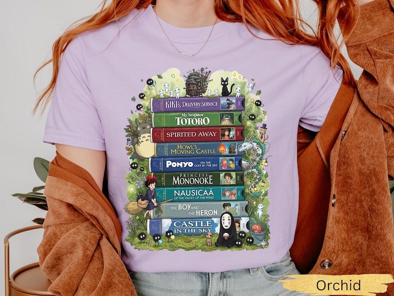 K&ouml;nnte beinhalten: Orchideenfarbenes T-Shirt mit einem Design aus bunten B&uuml;chern, darunter Titel wie "Kiki's Delivery Service" und "Mein Nachbar Totoro." Das Design enth&auml;lt Illustrationen von Charakteren und Elementen aus den Filmen, umgeben von skurrilen Details.