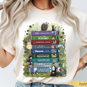 K&ouml;nnte beinhalten: Elfenbeinfarbenes T-Shirt mit einem Stapel bunter B&uuml;cher, darunter "Kiki's Delivery Service" und "Totoro." Das Design zeigt animierte Charaktere und einen gr&uuml;nen Bl&auml;tterrahmen. Das Shirt ist aus weichem Material.