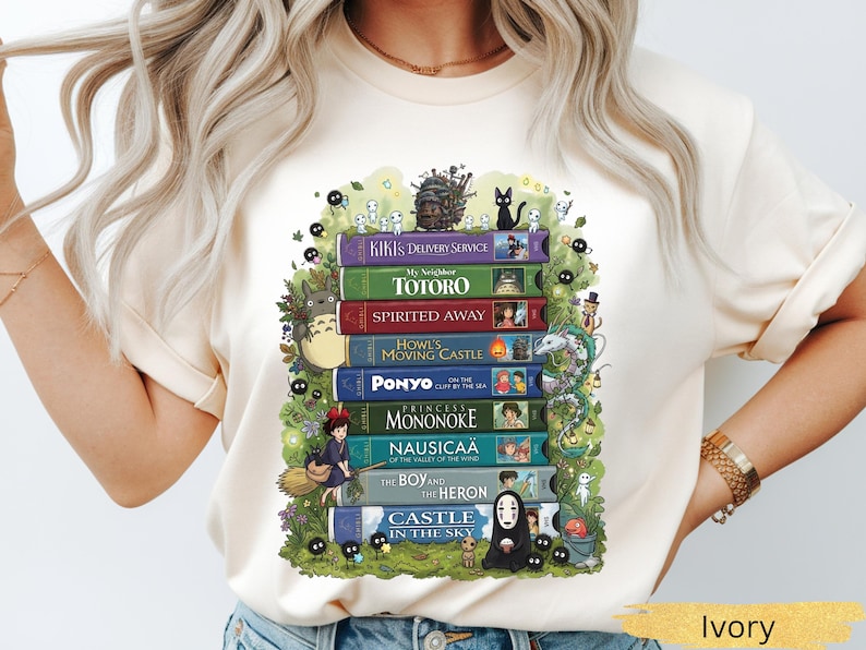 K&ouml;nnte beinhalten: Elfenbeinfarbenes T-Shirt mit einem Stapel bunter B&uuml;cher, darunter "Kiki's Delivery Service" und "Totoro." Das Design zeigt animierte Charaktere und einen gr&uuml;nen Bl&auml;tterrahmen. Das Shirt ist aus weichem Material.