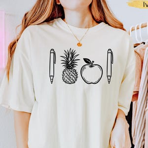 Op de afbeelding: Ivoor kleurig T-shirt met een zwarte lijntekening van twee pennen, een ananas en een appel. Het shirt heeft een losse pasvorm en korte mouwen. Het woord "Ivory" is goudkleurig gedrukt in de rechterbovenhoek.