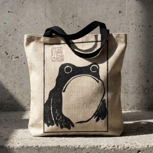 May include: A beige canvas tote bag featuring a black frog illustration. The frog is within a rectangular frame, with a Japanese stamp in the top left corner. The bag has black handles.