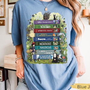 K&ouml;nnte beinhalten: Hellblaues T-Shirt mit einem farbenfrohen Grafikdesign von gestapelten B&uuml;chern mit Titeln wie "Totoro" und "Spirited Away". Das Design beinhaltet animierte Charaktere und Bl&auml;tter. Ein l&auml;ssiges, bequemes Kleidungsst&uuml;ck.