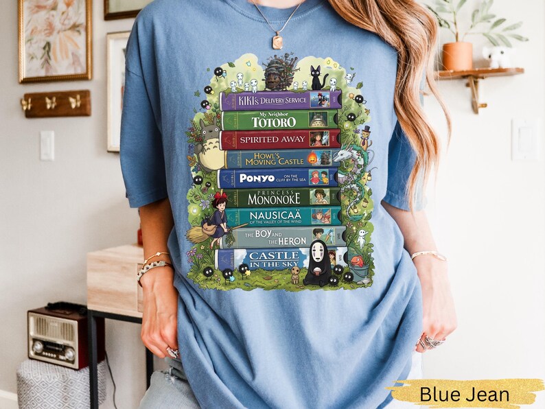 K&ouml;nnte beinhalten: Hellblaues T-Shirt mit einem farbenfrohen Grafikdesign von gestapelten B&uuml;chern mit Titeln wie "Totoro" und "Spirited Away". Das Design beinhaltet animierte Charaktere und Bl&auml;tter. Ein l&auml;ssiges, bequemes Kleidungsst&uuml;ck.