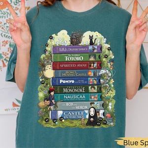 K&ouml;nnte beinhalten: Blau-Fichte-T-Shirt mit einem Design aus bunten B&uuml;chern, darunter "Kikis kleiner Lieferservice" und "Mein Nachbar Totoro." Die Illustrationen zeigen Charaktere und Szenen aus den Filmen. Das T-Shirt hat eine lockere Passform.