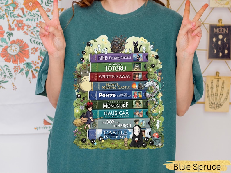 K&ouml;nnte beinhalten: Blau-Fichte-T-Shirt mit einem Design aus bunten B&uuml;chern, darunter "Kikis kleiner Lieferservice" und "Mein Nachbar Totoro." Die Illustrationen zeigen Charaktere und Szenen aus den Filmen. Das T-Shirt hat eine lockere Passform.
