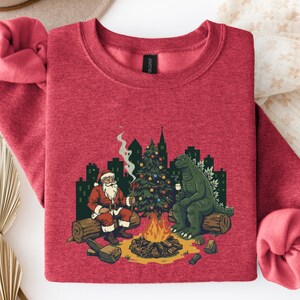 May include: Red sweatshirt featuring a graphic of Santa Claus and Godzilla by a campfire, with a Christmas tree and city skyline. Santa wears a red suit and hat. Godzilla is green, with a mug.