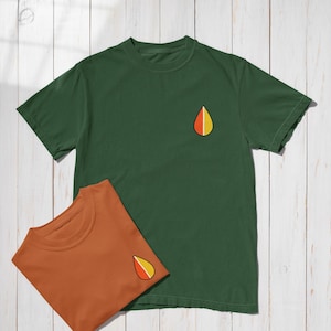 May include: Two short-sleeved t-shirts, one dark green and one rust-colored, are displayed on a white wooden surface. Each shirt features a small, embroidered teardrop-shaped design with red, yellow, and orange segments. The shirts are neatly folded.