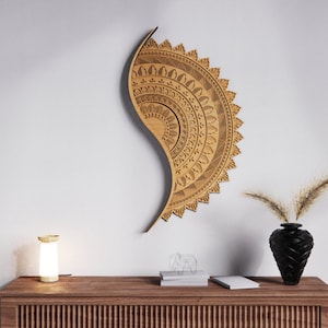 May include: A wooden wall art piece with a detailed mandala design is mounted on a white wall. The art is a light brown color and has a curved, semi-circular shape. A wooden cabinet with a lamp, books, and a black vase with dried plants is below the art.
