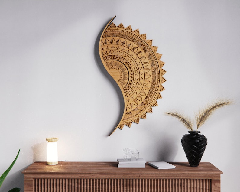 May include: A wooden wall art piece with a detailed mandala design is mounted on a white wall. The art is a light brown color and has a curved, semi-circular shape. A wooden cabinet with a lamp, books, and a black vase with dried plants is below the art.