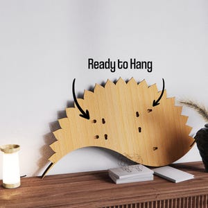 May include: A wooden wall decor piece with a sunburst design. The piece is light brown and has a scalloped edge. The text "Ready to Hang" is above the decor. A small lamp and a black vase with dried plants are on a wooden surface.