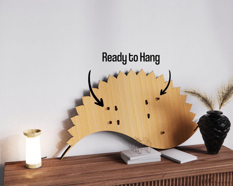 May include: A wooden wall decor piece with a sunburst design. The piece is light brown and has a scalloped edge. The text "Ready to Hang" is above the decor. A small lamp and a black vase with dried plants are on a wooden surface.