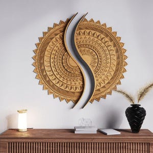 May include: A large, intricately carved wooden wall art piece in a sunburst design, split by a curved, light-colored element. The artwork is mounted on a white wall above a wooden cabinet with a lamp and a black vase.