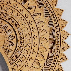 May include: Close-up of a wooden mandala with intricate, laser-cut patterns. The design features concentric circles with floral and geometric elements. The wood has a natural, light brown color, and the details are finely etched.