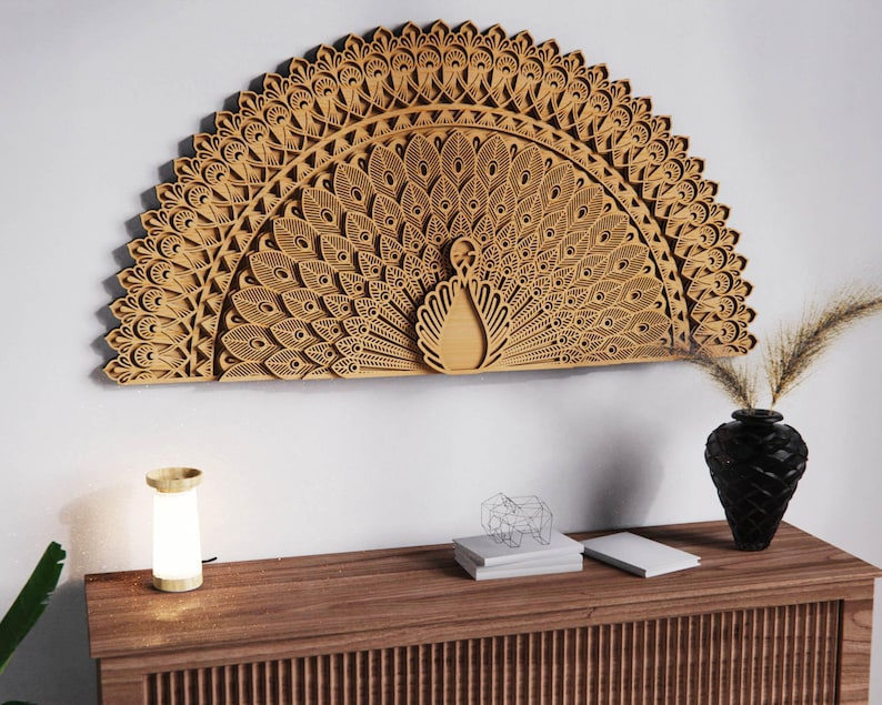 May include: A large, intricately carved wooden peacock wall art piece. The peacock's fanned tail is the focal point, with detailed feather patterns. The artwork is mounted on a white wall above a wooden cabinet with a lamp, vase, and decorative items.