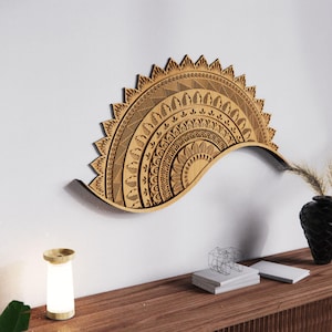 May include: A wooden, half-circle wall shelf with intricate mandala-style patterns. The shelf is a light brown color and has a curved design. It is mounted on a white wall above a wooden table with a black vase and a small lamp.