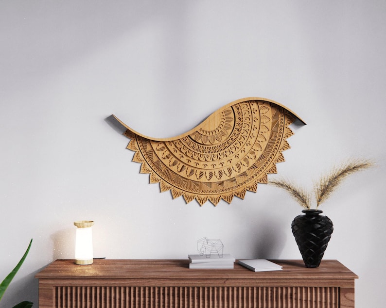 May include: A large, intricately carved wooden wall hanging in a wave shape, featuring detailed geometric patterns. The piece is a light brown color and is mounted on a white wall. A black vase with dried plants sits on a wooden cabinet below.