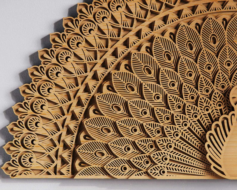 May include: A detailed wooden peacock feather design, laser-cut with intricate patterns. The artwork features a fan-shaped arrangement of feathers with eye-like details and geometric shapes. The natural wood color and layered construction create a 3D effect.