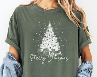May include: Moss green t-shirt with a white Christmas tree design and the words "Merry Christmas". The graphic includes a detailed tree, gifts, and stars. The shirt is a relaxed fit.