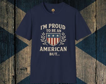 Puede incluir: Camiseta azul marino con el texto blanco "I'M PROUD TO BE AN AMERICAN BUT...". El diseño incluye un escudo con rayas rojas y blancas, estrellas azules y ramas de laurel. Cuello redondo.