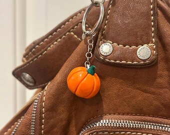 May include: A small, orange pumpkin charm with a green stem hangs from a silver keychain. The charm is attached to a brown leather handbag with silver hardware. The bag has visible stitching and a zipper.
