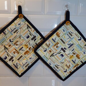 May include: Two square pot holders with a cream-colored background featuring various national park illustrations and text. The pot holders have a black border, a black loop, and a wooden button for hanging. The design includes bears, birds, and park names.
