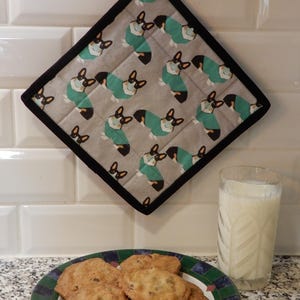 May include: A gray, square potholder with a black border features a repeating pattern of corgi dogs wearing teal sweaters. A plate of cookies and a glass of milk are in the foreground.
