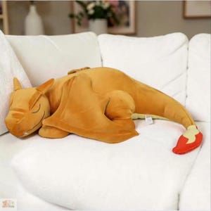 Könnte beinhalten: Ein orangefarbenes Charizard-Plüschkissen in entspannter Pose. Das Kissen hat einen detaillierten Kopf mit geschlossenen Augen, große Flügel und einen langen Schwanz mit roter und gelber Flammenspitze. Es liegt auf einer weißen Oberfläche.