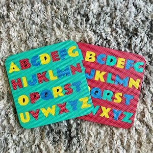 May include: Two colorful foam alphabet tiles. One is teal with yellow, red, blue, and green letters. The other is red with yellow, blue, and green letters. The tiles are on a textured, light-colored surface.