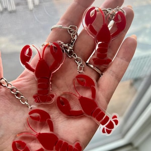 May include: Four red lobster-shaped keychains with silver key rings and chains. The lobsters are made of a clear material with red accents, and are held in a hand.