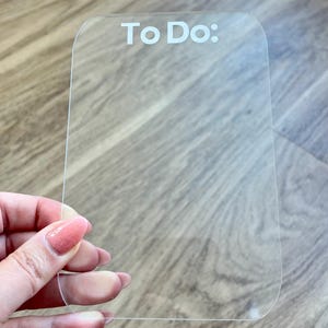 May include: A clear acrylic To Do list board with rounded corners. The words "To Do:" are printed in white at the top. The board is held by a hand with painted nails.