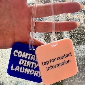 May include: Two square luggage tags. One is blue with the words "CONTAIN DIRTY LAUNDRY" in white. The other is peach with the words "tap for contact information" in black. Both have clear plastic loops.