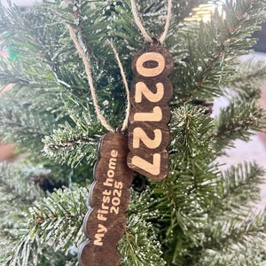 May include: Two wooden Christmas ornaments hanging on a tree. One ornament displays the year "2025" and the text "My first home". The other ornament displays the year "2027". Both are brown with a natural wood grain.