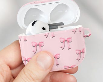 May include: Pink AirPods case with a bow pattern, held open to reveal white earbuds. One earbud is held separately. The case is pink with a repeating pattern of pink bows. The earbuds are white.