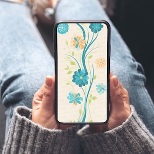 May include: A smartphone displaying a floral wallpaper design. The wallpaper features teal and orange flowers with green leaves on a cream background. The phone is held by a person wearing a gray sweater and blue jeans.