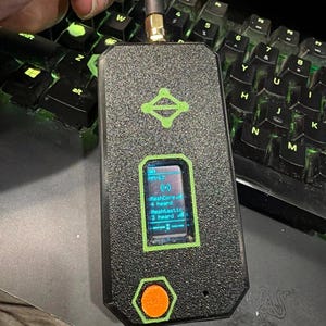 May include: A black textured handheld radio device with a green logo, a small digital screen displaying text including MeshCore and Meshtastic, a single orange circular button, and a vertical antenna extending from the top edge.