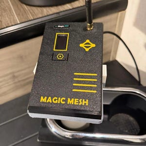 May include: A black rectangular electronic device with a textured finish, featuring the yellow text MAGIC MESH and Magic PTT. It includes a small vertical antenna, a rectangular screen, a yellow geometric icon, and four horizontal yellow lines.